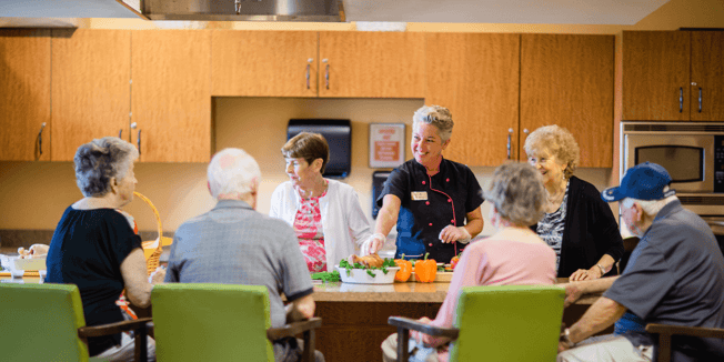 A group of elderly residents sitting in the kitchen of a senior living community while the chef prepares a chicken dinner. 