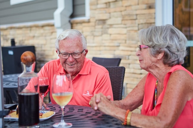 Two seniors drinking wine and eating at an outdoor dining table at a senior living community